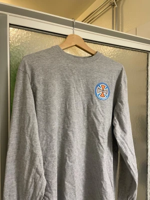 Grå långärmad t-shirt Independent - Grå långärmad t-shirt från Independent Truck Company med stort tryck på ryggen och mindre logga på bröstet. Klassisk passform och rund halsringning, perfekt för dig som gillar streetwear och skate.