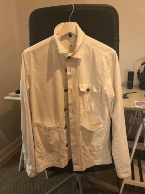 Vit overshirt från River Island - Stilren vit overshirt från River Island i kraftigt bomullstyg. Modellen har klassisk krage, knäppning framtill, en bröstficka med knapp och två stora framfickor. Perfekt lager på lager-plagg med avslappnad passform och snygga detaljer.