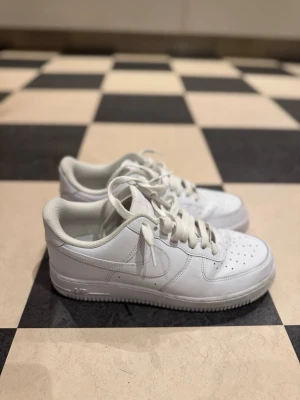 Vita Nike Air Force 1 sneakers - Klassiska vita Nike Air Force 1 sneakers i läder med perforerad tå, vita skosnören och låg profil. Ikonisk swoosh på sidan och bekväm sula som ger skön streetstil. Perfekta för dig som gillar tidlös och clean look. Använda 2 gånger