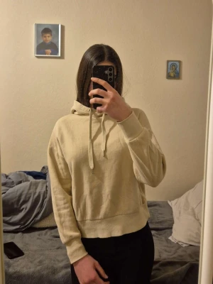 Beige hoodie med huva och dragsko - Snygg beige hoodie med huva och dragsko. Modellen är croppad med bred mudd nertill och långa ärmar. Perfekt för en avslappnad och trendig stil. Materialet ser ut att vara mjukt och bekvämt, troligen bomullsblandning. Inga synliga loggor eller tryck.