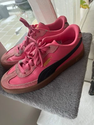 Rosa Puma sneakers med svart detalj - Snygga rosa sneakers från Puma med klassisk svart sidostrip och gummisula. Skorna har rosa mockadetaljer, matchande rosa snören och guldfärgad Puma-logga på sidan. Perfekta för dig som vill sticka ut med färg och sportig stil.