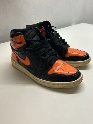 Nike Air Jordan 1 Shattered Backboard - Säljer ett par Nike Air Jordan 1 Shattered Backboard med svart och orange patentläder, klassisk high-top siluett och vita sulor. Ikonisk Swoosh på sidan och Air Jordan-logga vid ankeln. Grymma sneakers för dig som vill sticka ut.