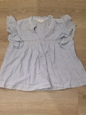 Ljusblå volangblus med spetsdetaljer - Supersöt ljusblå blus med volanger på axlarna och fina spetsdetaljer över bröstet. Blusen har en avslappnad passform och V-ringning. Tillverkad i ett tunt, luftigt tyg som känns perfekt för varmare dagar. Den är använd en gång och är fortfarande I jätte bra skick o inga fläckar. Storlek är mellan 158 till 164 kommer ifrån ginatricot 