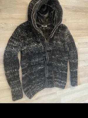 Mörkgrå stickad cardigan  med luva - Mysig mörkgråmelerad stickad kofta med luva och knappar framtill. Koftan har långa ärmar och är fodrad med mjukt, fluffigt material i luvan för extra komfort. Perfekt för kyliga dagar och en avslappnad stil.