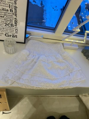 Vit volangkjol från Gina Tricot - Supersöt vit kortkjol från Gina Tricot med volanger och smock i midjan. Kjolen har ett diskret mönster och är perfekt för dig som vill ha en luftig och trendig look. Passar perfekt till sommaren och är lätt att matcha med olika toppar. Storlek 146-152 