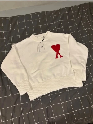 Vit stickad tröja från Ami Paris - Vit stickad tröja från Ami Paris med röd hjärtlogga och bokstaven A på bröstet. Tröjan har rund hals, ribbade muddar och en avslappnad passform. Perfekt för dig som gillar stilrena plagg med en twist.