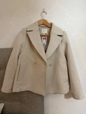 Beige kappa med bred krage från H&M - Stilren beige kappa från H&M med bred krage och dubbelknäppning framtill. Kappan har två raka fickor och är tillverkad i ett mjukt, strukturerat tyg. Perfekt för dig som vill ha en klassisk och enkel look.