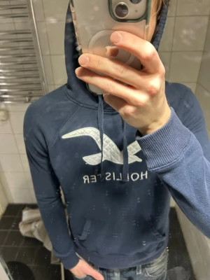 Blå hoodie från Hollister - Mörkblå hoodie från Hollister med stor vit logga och text på bröstet. Klassisk huva med dragsko och magficka framtill. Tillverkad i mjukt material och har ribbade muddar vid ärmslut och nederkant. Perfekt för en avslappnad och stilren look.