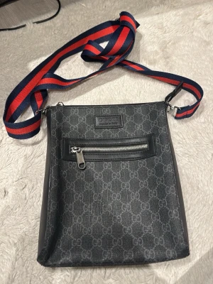 Svart axelväska från Gucci - Snygg svart axelväska från Gucci med klassiskt GG-mönster och en framficka med dragkedja. Väskan har en bred axelrem i rött och blått, samt detaljer i läder och metall. Perfekt för dig som vill ha en stilren och trendig accessoar.