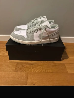 Nike Air Jordan 1 Low *sällsynta* - Snygga Air Jordan 1 Low sneakers i vitt och mintfärg som inte går att få tag på i Sverige nu mera. Ovandelen är i slät vit läder med grå mockadetaljer på sidor, häl och snörning. Klassisk Jumpman-logga på plösen och perforerad tå för extra ventilation. Perfekta för dig som gillar stilrena och sportiga sneakers.