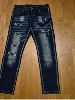 Mörkblå distressed jeans Dsquared2 - Jag säljer ett par original Dsquared2-jeans i mycket fint skick. Jeansen är i storlek 48 enligt italiensk storleksskrivning, vilket motsvarar cirka W33. De har en snygg och modern design med slitningar och diskreta färgstänk som är en del av originalutförandet. Passformen är slim och sitter bra på kroppen. Jeansen är väl omhändertagna och har inga oavsiktliga skador. Först till kvarn gäller och pris kan diskuteras vid snabb och smidig affär