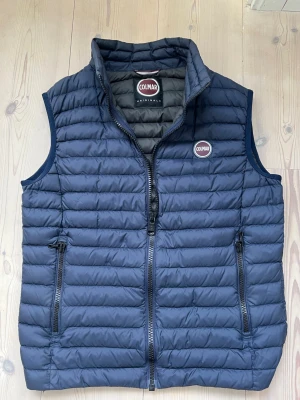 Mörkblå dunväst från Colmar - Snygg mörkblå dunväst från Colmar med quiltad design och hög krage. Västen har två dragkedjeförsedda fickor och ett tydligt Colmar-märke på bröstet. Perfekt att slänga över en hoodie eller tröja för extra värme och stil🤩. Skick 9/10🙌. Tveka inte att ställa frågor🤝