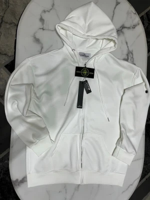 Stone Island vit zip hoodie – ny med tags - Säljer en Stone Island zip hoodie i vit färg. Plagget är helt nytt med tags kvar och aldrig använt.  Storlek: M Passform: Normal fit  Kan skicka fler bilder och detaljer vid intresse.