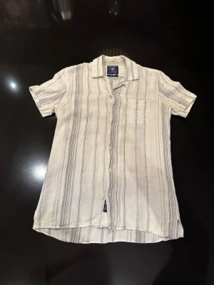 Randig skjorta från Robbie Moor - Snygg kortärmad skjorta från Robbie Moor i ljusbeige med tunna svarta ränder. Skjortan har klassisk krage, bröstficka och knäppning framtill. Perfekt för en avslappnad och stilren look. Lägg märke till slitage vid en av knapparna.