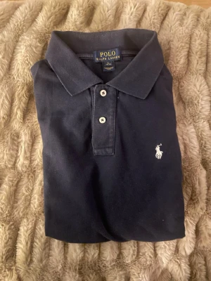 Svart pikétröja från Ralph Lauren - Klassisk mörkblå pikétröja från Ralph Lauren med vit broderad logga på bröstet. Tröjan har korta ärmar, krage och knappar framtill. Tillverkad i mjuk bomull som känns skön mot huden. Perfekt för en stilren och avslappnad look. Storlek L i barn storlek 14-16 år. Mycket bra skick.