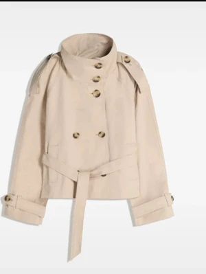 Kort beige trenchcoat med bälte - Stilren kort trenchcoat i beige med dubbelknäppning, höga axelklaffar och knytskärp i midjan. Jackan har stora knappar i kontrastfärg och justerbara ärmslut. Tillverkad i ett slätt tyg som ger en klassisk look. Helt ny och ej andvänd, dock tyckte jag av prislappen för snabbt🥰
