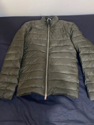 Olivgrön dunjacka med dragkedja - Snygg olivgrön dunjacka med hög krage och hel dragkedja framtill. Jackan har ett quiltat mönster med horisontella sömmar och är lätt vadderad för extra värme. Perfekt för kalla vinterdagar och enkel att matcha med olika outfits. Ganska stilig jacka och udda  pris går själv klart att lösa snabbare köp bättre pris 