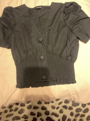 Svart topp med puffärm från Gina Tricot - Snygg svart topp från Gina Tricot med puffärmar och bred smock i midjan. Toppen har v-ringning, stora dekorativa knappar framtill och kortare passform. Perfekt att styla med höga jeans eller kjol. Den är mörkare i verkligheten❤️