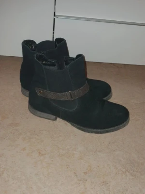 Svarta mockastövletter från rieker  - Snygga svarta stövletter i mocka-look med dekorativ rem- och knappdetalj på sidan. Klassisk rund tå och låg klack gör dem perfekta till jeans eller kjol. Dragkedja på insidan för enkel påtagning. Stilrena och passar till många outfits.