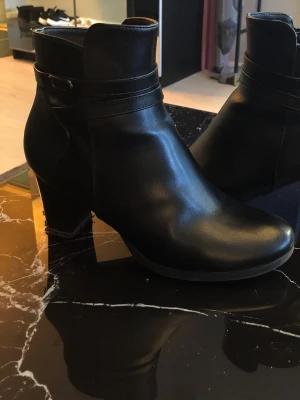 Svarta skinnstövletter med klack  - Snygga svarta stövletter i slätt skinn med rundad tå och stabil klack. Dekorativ rem med metallspänne runt ankeln ger en stilren look. Perfekta till jeans eller kjol för en trendig outfit. Nypris 599. Strl 41 men passar strl 38-41