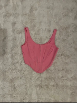 Rosa korsettopp med dragkedja - Snygg rosa korsettopp med breda axelband och markerad midja. Toppen har en dragkedja framtill och är croppad med en spetsig nederkant. Perfekt för att lyfta vilken outfit som helst! Toppen är köpt på en butik i Malta och har tyvärr ingen lapp.