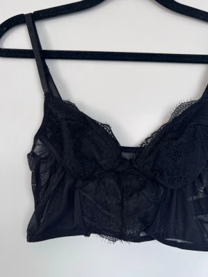 Svart spetsbralette med tunna axelband, storlek S - Snygg svart bralette i transparent spets med tunna justerbara axelband. Mjuk och bekväm modell utan bygel, perfekt för dig som gillar en lättare känsla. Spetsdetaljer framtill och mesh på sidorna ger en trendig look.