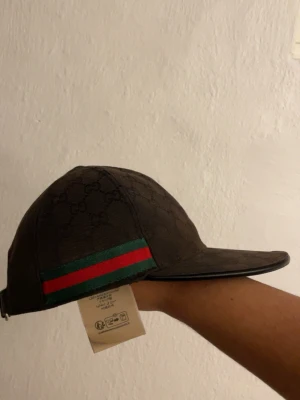 Gucci keps med GG-mönster - Snygg keps från Gucci med klassiskt GG-monogram över hela kepsen. På sidan finns en grön och röd rand som ger den där ikoniska Gucci-looken. Justerbar rem baktill för perfekt passform. Tillverkad i slitstarkt tyg som känns lyxigt. (Lite missfärgad/solbränd) Pris går att diskutera. (Byten funkar också🙌🏽)