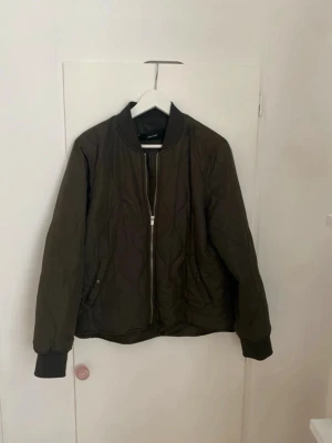 Olivgrön quiltad bomberjacka Vero Moda - Snygg olivgrön bomberjacka från Vero Moda med quiltat mönster, dragkedja framtill och ribbade muddar vid ärmslut och krage. Jackan har två fickor med knappstängning och en klassisk bomberjacke-look. Perfekt för dig som vill ha en stilren och trendig jacka.