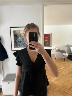 Svart omlottblus med volangärm - Snygg svart omlottblus med korta volangärmar och v-ringning. Blusen har broderade detaljer och en feminin passform som ger en trendig look. Perfekt att styla med jeans eller kjol.