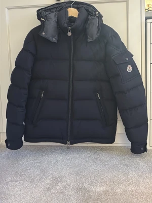 Svart dunjacka från Moncler - Svart dunjacka från Moncler med avtagbar huva och två dragkedjefickor framtill. Ordinarie pris 23 000 kr. Knappast använd! Jackan är Moncler Man. 