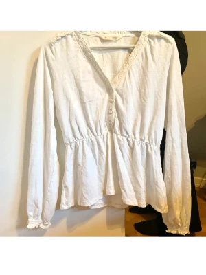 Vit blus med spets från Odd Molly - Söt vit blus från Odd Molly med v-ringning och spetsdetaljer längs halsringningen. Blusen har knappar framtill, lång ärm med resår vid ärmslut och en lätt rynkad midja som ger en feminin siluett. Tillverkad i mjukt bomullstyg.
