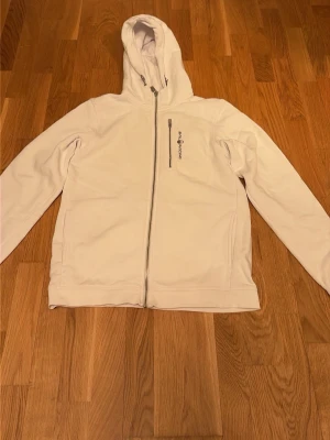 Vit Zip Hoodie från Sail Racing - Snygg vit hoodie från Sail Racing med dragkedja och klassisk logga broderad på bröstet. Tröjan har huva med snörning och två stora fickor framtill, samt en ficka med dragkedja vid bröstet. Perfekt för en clean och stilren look.                                           