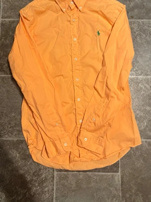 Orange skjorta från Ralph Lauren - Snygg orange skjorta från Ralph Lauren med klassisk button-down krage och grönt broderat logotyp på bröstet. Skjortan har långa ärmar och knäppning framtill. Perfekt för en stilren och avslappnad look.