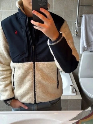  Polo Ralph Lauren jacka - Mysig teddyjacka i beige med svarta detaljer och dragkedja framtill. Jackan har hög krage, två fickor med dragkedja och Polo Ralph Laurens röda logotyp på bröstet. Perfekt för kalla dagar och en cool look.