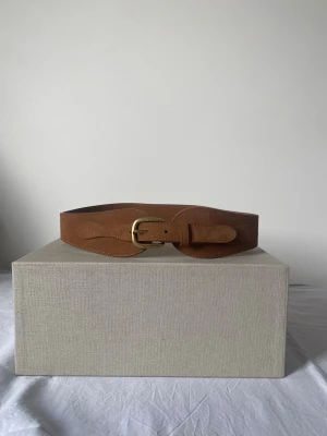Brunt brett mockabälte med mässingsspänne - Snyggt brunt bälte i mocka med brett, formsytt band och ett klassiskt mässingsfärgat spänne. Perfekt för att lyfta vilken outfit som helst med en vintagekänsla. Bältet har mjuka former och synliga sömmar för extra detalj.