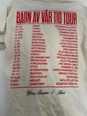 Barn av vår tid tour t-shirt - Vit t-shirt med röd text på ryggen som listar turnédatum och städer för 'Barn av vår tid tour'. T-shirten har klassisk passform och korta ärmar. Materialet känns som bomull och trycket är tydligt och stort.