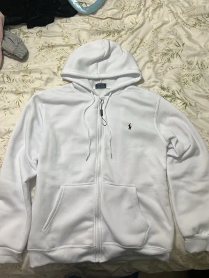 Vit zip hoodie från Polo Ralph Lauren - Snygg vit hoodie med dragkedja från Polo Ralph Lauren. Klassisk design med huva och två fickor framtill. Ikoniska Polo-loggan broderad på bröstet i svart. Perfekt för en clean och stilren look. Tillverkad i mjukt bomullsmaterial för extra komfort.