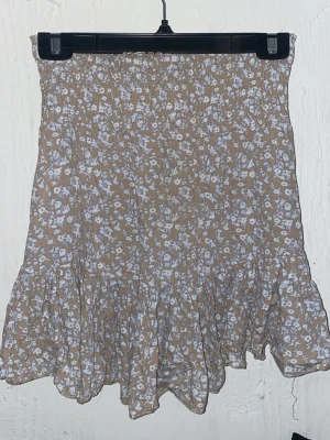 Beige blommig volangkjol - Söt kortkjol i beige med vitt blommönster i storlek M men insydd i midjan till S. Ifrån Gina tricot. Kjolen är gjord i ett lätt och luftigt material, perfekt för varmare dagar. Passar till både sneakers och sandaler.