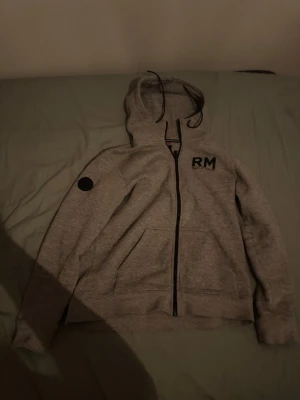 Grå hoodie från Race Marine - Snygg grå hoodie från Race Marine med svart RM-tryck på bröstet och dragkedja framtill. Hoodien har justerbar huva med snören och mjukt fleecefoder på insidan. Perfekt för en avslappnad och sportig stil.