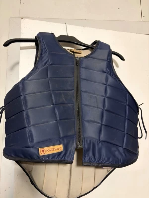 Mörkblå skyddsväst från Racesafe - Mörkblå skyddsväst från Racesafe med dragkedja framtill och vadderad design för extra säkerhet. Västen har justerbara snören i sidorna 