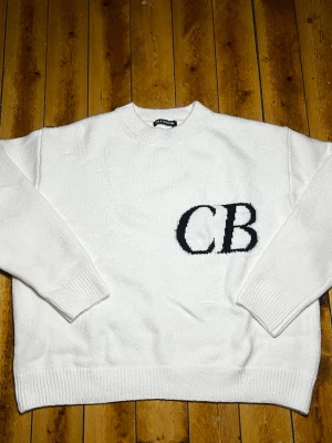 Vit stickad tröja Cole Buxton - Säljer en vit stickad tröja från Cole Buxton med svarta bokstäver 'CB' på bröstet. Tröjan har rund hals, långa ärmar och ribbade muddar vid ärmslut och nederkant. Perfekt för dig som gillar enkel men snygg stil.