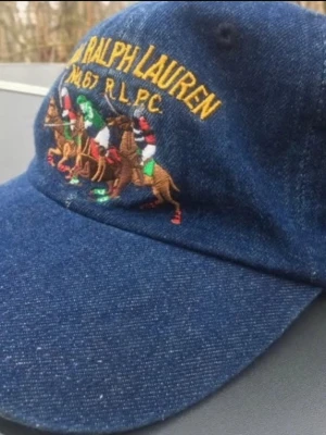 Blå denimkeps från Polo Ralph Lauren - Snygg blå denimkeps från Polo Ralph Lauren med broderat polomotiv och gul logga framtill. Kepsen är i 100% bomull och har justerbar passform. Perfekt accessoar för en sportig och avslappnad stil.
