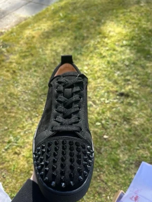 Svarta sneakers med nitar från Louboutin - Säljer ett par svarta sneakers från Christian Louboutin med mattsvarta nitar på tån. Skorna har ovandel i mocka och läder, svarta skosnören och en klassisk låg siluett. Perfekta för dig som vill sticka ut med en edgy stil.