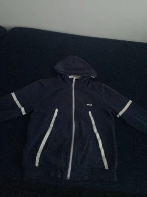 Mörkblå BOSS tröja med dragkedja  - Snygg mörkblå hoodie från BOSS med vit dragkedja och vita detaljer längs fickorna och ärmarna. Klassisk huva och diskret BOSS-logga på bröstet. Perfekt för en avslappnad och stilren look.
