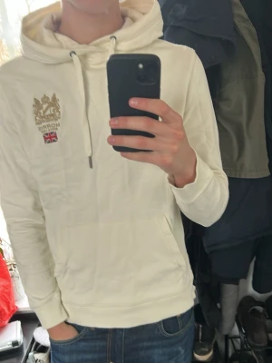 Morris hoodie - Säljer en stilren vit/creme hoodie från Morris med broderad logga och brittisk flagga på bröstet. Tröjan har en klassisk känguruficka och justerbar huva med snören. Perfekt för en avslappnad stil. Liten fläck på höger armen men syns knappt vid användning(bild 2)