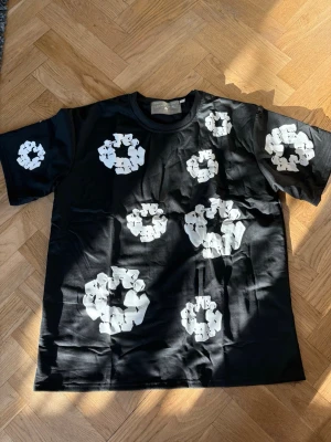 Svart t-shirt med blommor från Denim Tears - Svart t-shirt från Denim Tears med vita blommiga tryck över hela framsidan och på ärmarna. Klassisk passform med rund halsringning och korta ärmar. Perfekt statement-plagg för dig som vill sticka ut.