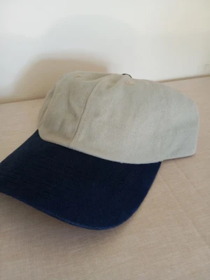 Beige och marinblå keps - Snygg keps med beige krona och marinblå skärm. Klassisk sexpanel-design med böjd skärm och justerbar rem bak. Tillverkad i bomullstyg som ger en avslappnad och stilren look.