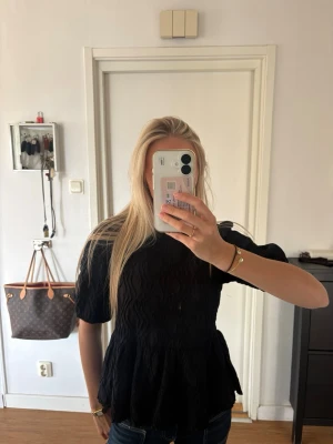Svart puffärmad blus med öppen rygg - Trendig svart blus med puffiga korta ärmar och strukturerat tyg. Blusen har en snygg öppen rygg med knytband upptill och en lätt utsvängd midja. Perfekt för dig som vill sticka ut med en cool och modern look.