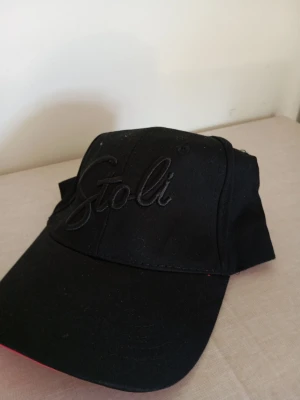 Svart keps med Stoli-brodyr - Snygg svart keps med böjd skärm och broderad Stoli-logga framtill. Klassisk modell med justerbar rem bak. Tillverkad i bomullstyg för en bekväm känsla. Perfekt för en avslappnad streetwear-look.