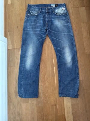 Blå raka jeans från Replay - Säljer ett par blå jeans från Replay med raka ben och slitna detaljer på fickor och benslut. Jeansen har klassisk femficksdesign, ljusa tvättade partier och tydliga sömmar. Perfekta för en avslappnad och trendig look. Har litet hål i bakfickan som syns på andra bilden